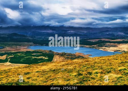 Una vista panoramica di un lago incastonato tra colline, con un cielo blu e una spettacolare catena montuosa in lontananza, coperta da nuvole. Foto Stock