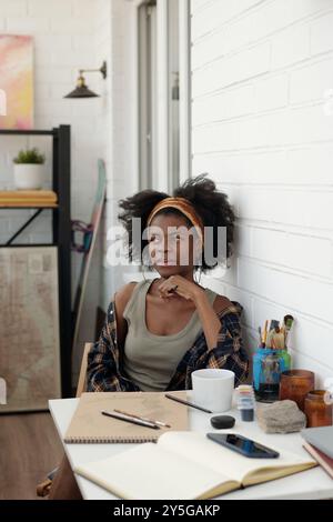 Donna afro-americana che contempla con cura idee creative in un ambiente elegante e ben illuminato in studio con opere d'arte e piante sullo sfondo, circondato da forniture di pittura e schizzi Foto Stock