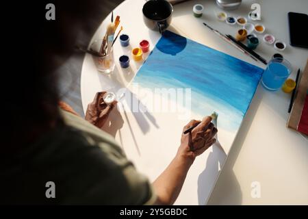 Persona che dipinge tonalità blu su tela tra le forniture d'arte, tra cui pennelli, contenitori di pittura e un bicchiere d'acqua, creando arte astratta con tratti morbidi e colori di fusione Foto Stock