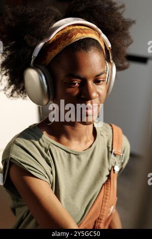 Donna afroamericana sorridente mentre indossa le cuffie, ascoltando musica in un ambiente interno moderno. Capelli legati all'archetto, mentre i raggi del sole illuminano lo spazio circostante Foto Stock
