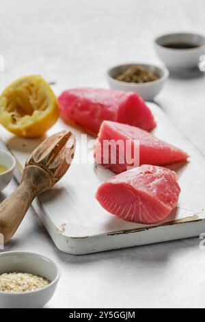 Tre vivaci bistecche di tonno riposano su un tagliere vicino alle metà del limone e alle piccole ciotole ripiene di condimento. L'allestimento suggerisce un piatto saporito nel Foto Stock