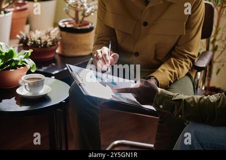 Due professionisti si sono impegnati nella revisione e nella discussione dei documenti finanziari in un ambiente accogliente e pieno di impianti che presenta piante da interno, tazza di caffè su un tavolo di legno durante una riunione di lavoro Foto Stock