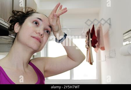 La casalinga bruna brasiliana sembra stanca in una piccola lavanderia. Donna bianca espressiva che fa i lavori di casa. Governante caucasica. Foto Stock