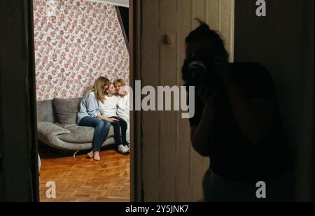 Tra la carta da parati floreale, due amici condividono un momento di gioia su un divano, mentre una terza persona cattura la loro felicità nella calda luce della sera. Foto Stock