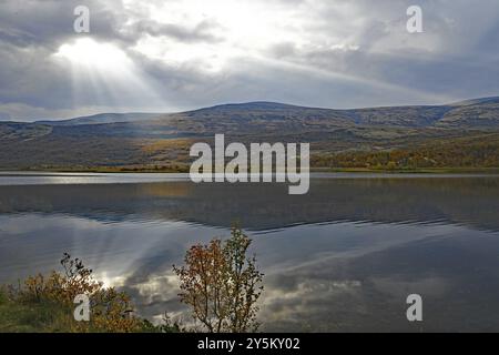 Un lago calmo con montagne sullo sfondo, raggi di sole che si infrangono tra le nuvole, colori autunnali dominano il paesaggio, Valasjoen, Hjerkinn, Dovref Foto Stock