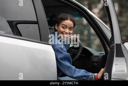 Amichevole sorridente millennial afroamericana driver femminile apre le porte e. cerca il compagno di viaggio Foto Stock