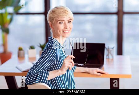 Donna d'affari sorridente che posa tenendo gli occhiali seduti in ufficio Foto Stock