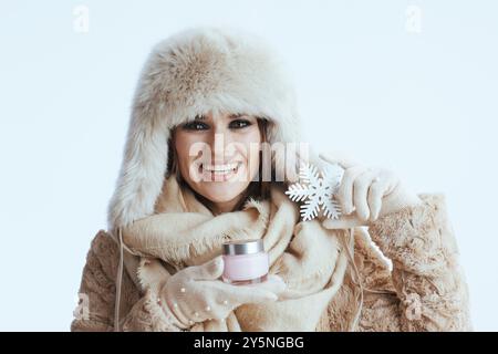 donna felice ed elegante con cappotto invernale e cappello in pelliccia isolato su sfondo bianco con guanti bianchi con crema viso e fiocco di neve. Foto Stock