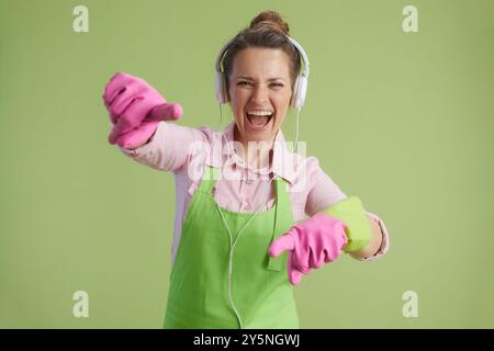 Pulizia di primavera. donna di mezza età moderna sorridente con grembiule verde e guanti di gomma su sfondo verde con cuffie rivolte verso la fotocamera. Foto Stock