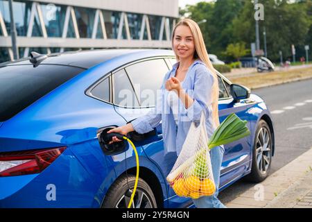 Una donna bionda sta accanto al suo veicolo elettrico blu, tenendo in mano una borsa di frutta e verdura mentre si carica Foto Stock