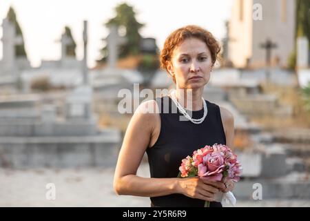 Donna in abito nero con bouquet di fiori è venuta al cimitero per onorare la memoria della persona amata defunta Foto Stock