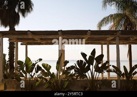 Un semplice rifugio in legno sul mare con pali rustici, incorniciati da piante tropicali. Il mare calmo è visibile sullo sfondo sotto un cielo limpido. Foto Stock
