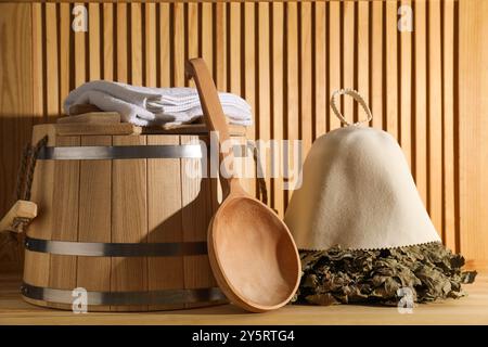 Frusta per sauna in quercia, cappello in lana di feltro, asciugamani, secchio e mestolo su una superficie in legno Foto Stock