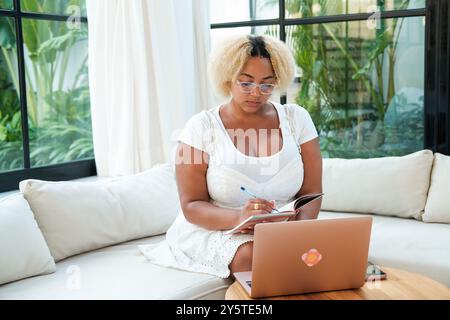 Donna afro-americana curvy con occhiali seduti in soggiorno e che lavora Foto Stock