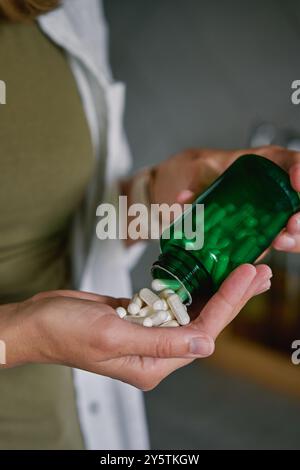 Primo piano di una donna che versa capsule bianche dal biberon verde alla mano. La persona sta assumendo pillole o vitamine. Concetto di integratori, medicina o salute c Foto Stock