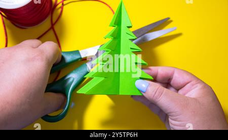 Donna che realizza oggetti di artigianato per decorazioni natalizie su sfondo giallo con filo decorativo natalizio e forbici Foto Stock