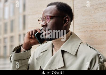 Uomo afroamericano sorridente che tiene in mano lo smartphone e parla in un ambiente urbano indossando un cappotto e occhiali eleganti Foto Stock