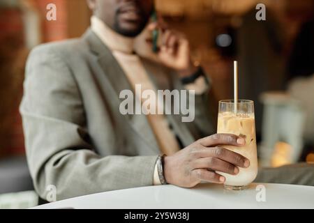 Uomo sfocato in tuta che tiene il telefono all'orecchio mentre prende il caffè ghiacciato con la cannuccia. Concentrarsi sulle bevande, con la persona leggermente offuscata sullo sfondo Foto Stock