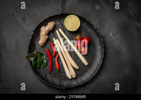 Ingredienti per preparare Tom Yum Kung. Citronella, peperoncino, zenzero, galangal, lime, coriandolo e pomodori ciliegini su un piatto nero contro una ba scura Foto Stock