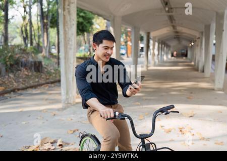 Young Man in bicicletta nel parco mentre utilizza lo smartphone sotto la passerella coperta in un giorno di sole Foto Stock