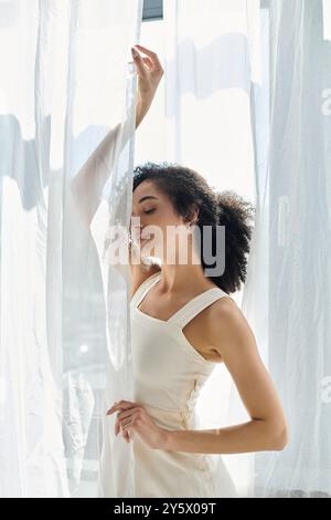 Una donna alla moda sta in piedi con tende che scorrono, immergendosi nella calda luce del sole mentre posa elegantemente. Foto Stock