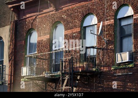 La luce del sole bagna la facciata di un edificio in mattoni d'epoca con finestre ad arco e un piccolo balcone adornato con una parabola satellitare. Foto Stock