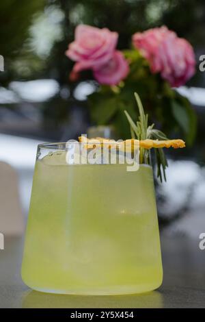 Un rinfrescante cocktail verde guarnito con una fetta di ananas essiccato e rosmarino, con rose rosa sullo sfondo. Foto Stock