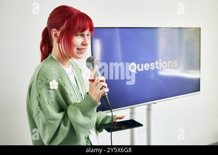 Giovane donna con capelli rossi che dà presentazione tenendo in mano microfono e telefono davanti allo schermo che mostra domande Foto Stock