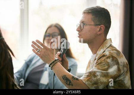 Giovane con gli occhiali che parla al microfono durante l'evento della conferenza con partecipanti sfocati in sottofondo. Indossa un abbigliamento casual e sfoggia un'espressione accattivante del viso Foto Stock