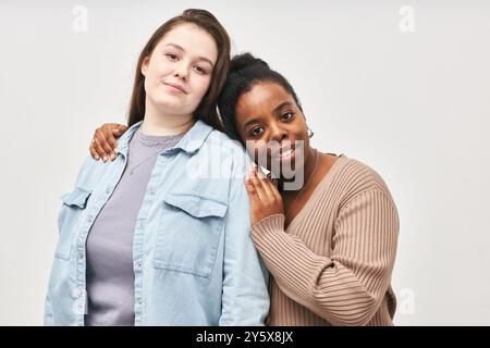 Due amici che si uniscono mentre si abbracciano indossando un abbigliamento casual e sorridenti. Lo sfondo è semplice e minimale, concentrandosi sul momento autentico condiviso tra di loro Foto Stock