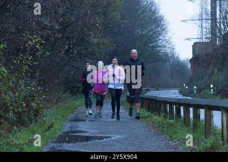 Gruppo di corridori che si allenano su un sentiero bagnato accanto a una recinzione di legno in una giornata nuvolosa. Foto Stock