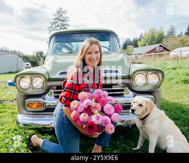 Donna sorridente con un bouquet di fiori seduto su un camion d'epoca con un cane in campagna. Foto Stock