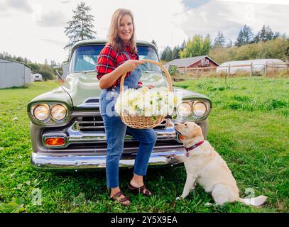 Una donna sorridente in una camicia a quadri con un cesto di fiori si erge accanto a un'auto classica con un labrador retriever giallo seduto accanto a lei in un campo erboso. Foto Stock