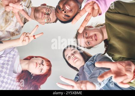 Gruppo di cinque amici che si formano in cerchio sorridendo e mostrando segni di pace. Catturato dal basso, creando un'atmosfera gioiosa e dinamica che celebra l'unità e la diversità Foto Stock