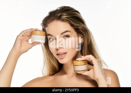 Questa bella giovane donna contiene due vasetti di prodotti per la cura della pelle, sorridendo elegantemente in una luce brillante. Foto Stock