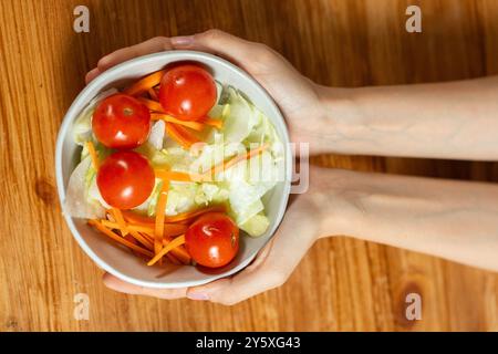 Vista dall'alto delle mani che reggono una ciotola bianca ripiena di insalata fresca, tra cui pomodori ciliegini, lattuga e carote tritate. L'immagine ritrae mangiato sano Foto Stock