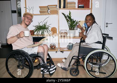 Amici sorridenti seduti su sedie a rotelle tenendo le piante e bevendo un caffè in un accogliente soggiorno con scaffali e sfondo verde, creando un'atmosfera calda e accogliente Foto Stock