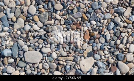 La spiaggia presenta una variegata collezione di ciottoli lisci e colorati sparsi lungo la riva, riflettendo la tranquillità della giornata Foto Stock