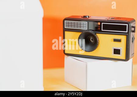 Una fotocamera vintage dal design giallo e nero si trova elegantemente su una scatola bianca, su uno sfondo arancione dinamico, evocando nostalgia e creatività Foto Stock