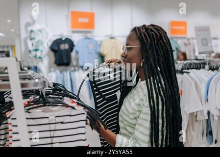 Una donna afroamericana sfoglia i top a righe in un negozio di abbigliamento, selezionando con cura gli articoli da acquistare in un viaggio di shopping informale. Foto Stock