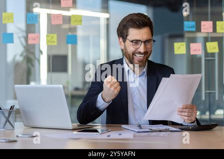 Professionista professionale in tuta che mostra entusiasmo durante la revisione dei documenti presso la workstation. L'uomo sorridente che lavora con un notebook in un ambiente open Office dimostra successo e motivazione. Foto Stock