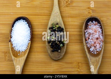 Tre cucchiai di legno disposti su un tagliere di bambù. Cucchiai ripieni di sale pretzel, grani di pepe interi e sale himalayano. Foto Stock