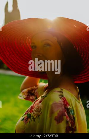 Ritratto di una donna che indossa un grande cappello rosso durante l'ora d'oro. La luce calda e soffusa del sole mette in risalto i bordi del cappello e proietta una splendida luce Foto Stock