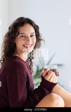 Donna sorridente che tiene in mano un bicchiere di tè, godendosi un momento di relax a casa Foto Stock