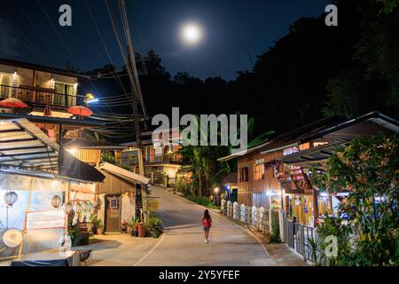 Una donna cammina lungo una strada del villaggio di Mae Kampong di notte, con una luna piena nel cielo. La strada è fiancheggiata da piccole case e aziende, northe Foto Stock