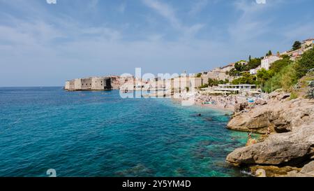 I visitatori si crogiolano al sole lungo la splendida spiaggia di Dubrovnik, Croazia, dove le acque turchesi si incontrano con le vivaci scogliere e le strutture storiche, creando una pittoresca fuga costiera per tutti. Foto Stock