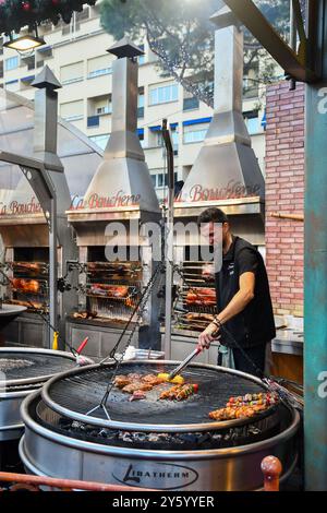 Un giovane cuoco sorridente che grigia spiedini di carne e pannocchie di mais al mercatino di Natale di Port Hercule, durante le vacanze invernali, Monte Carlo, Monaco Foto Stock