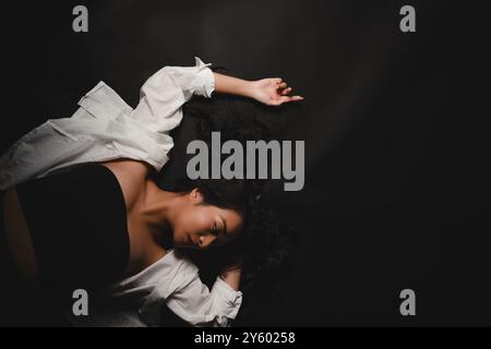 Una donna che indossa un top nero e una camicia bianca è sdraiata su un tessuto nero, con il viso inclinato lateralmente Foto Stock