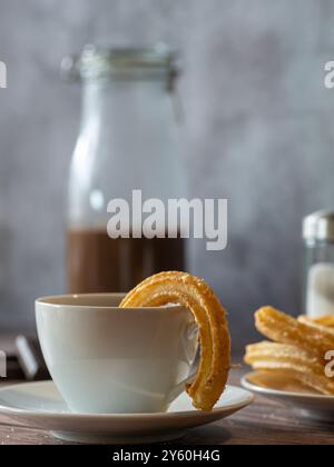 Primo piano di una tazza di cioccolata calda con un churro all'interno. Churros e bottiglia di cioccolato sullo sfondo Foto Stock