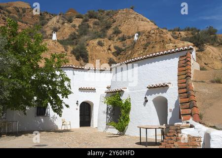 Edificio bianco con pareti di roccia, alberi e mobili rustici da esterno sotto un cielo azzurro soleggiato, hotel nelle grotte Pedro Antonio de Alarcon, Guadix, provincia di Granada Foto Stock